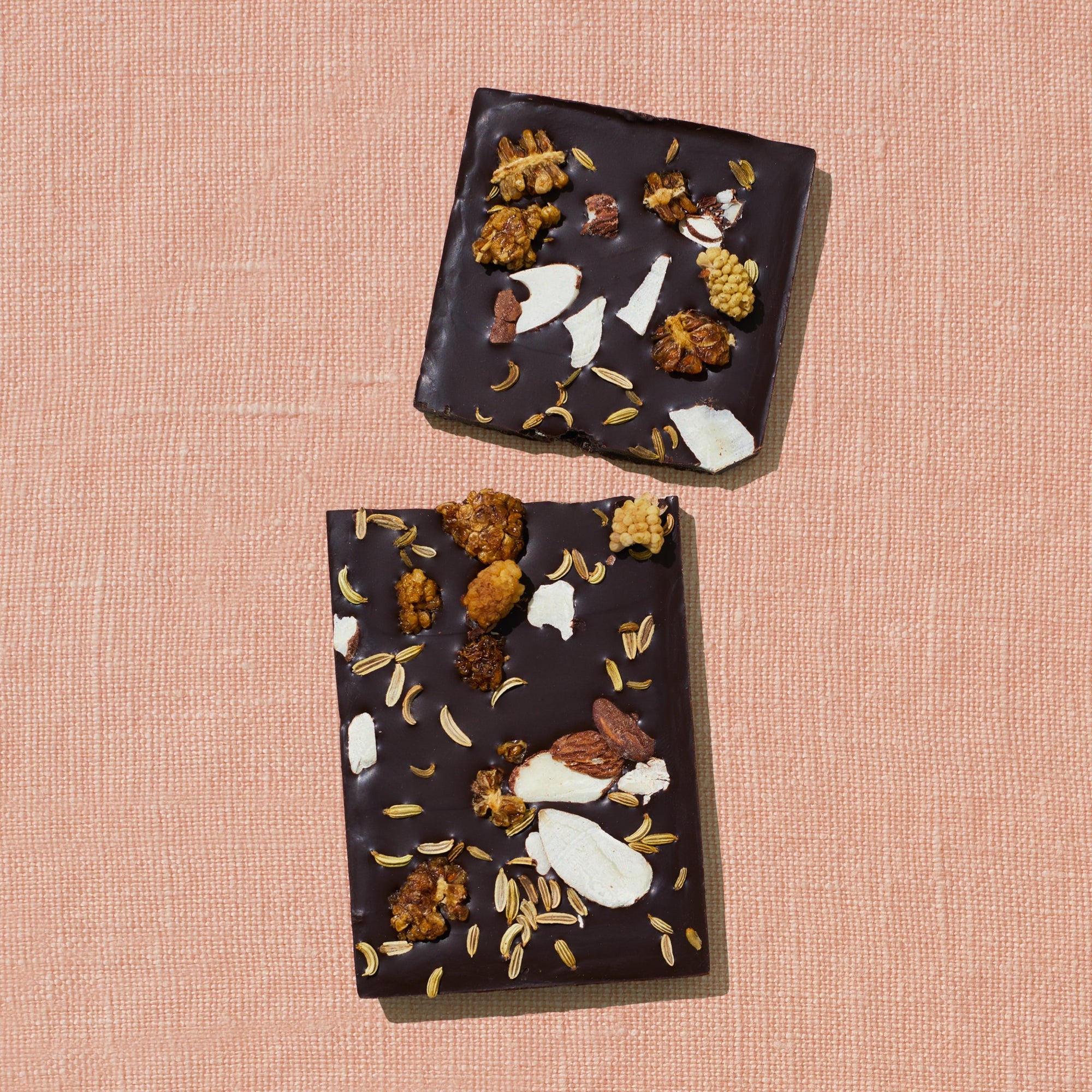 One chocolate bar that is topped with mulberries, almond flakes, and fennel is laid out on a warm brown background broken in two pieces.