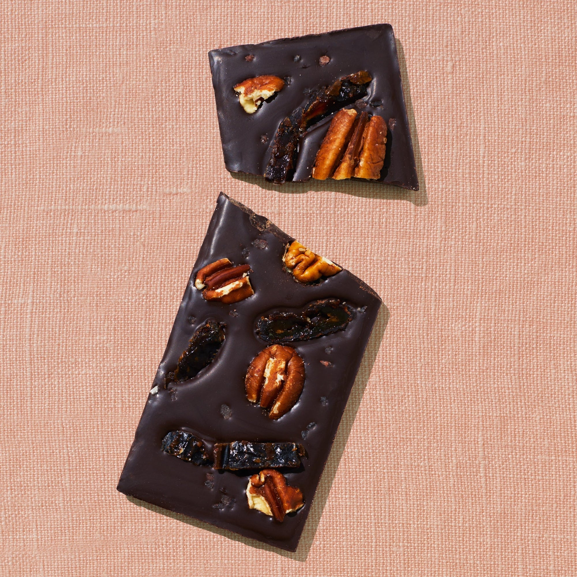 One chocolate bar that is topped with dates, pecans, and rock salt is laid out on a warm brown background broken in two pieces.