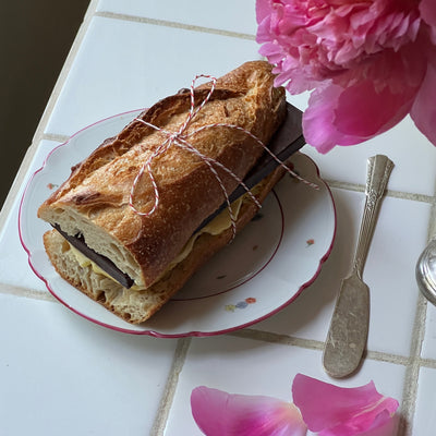 Sitting on top of a tiled background Spring & Mulberry Pure Dark Chocolate bar and butter in a crusty baguette, tied with a bow and surrounded by flowers and petals
