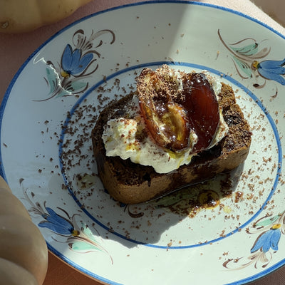 Slice of chocolate pumpkin bread topped with cream, dates, olive oil and chocolate shavings on a floral plate