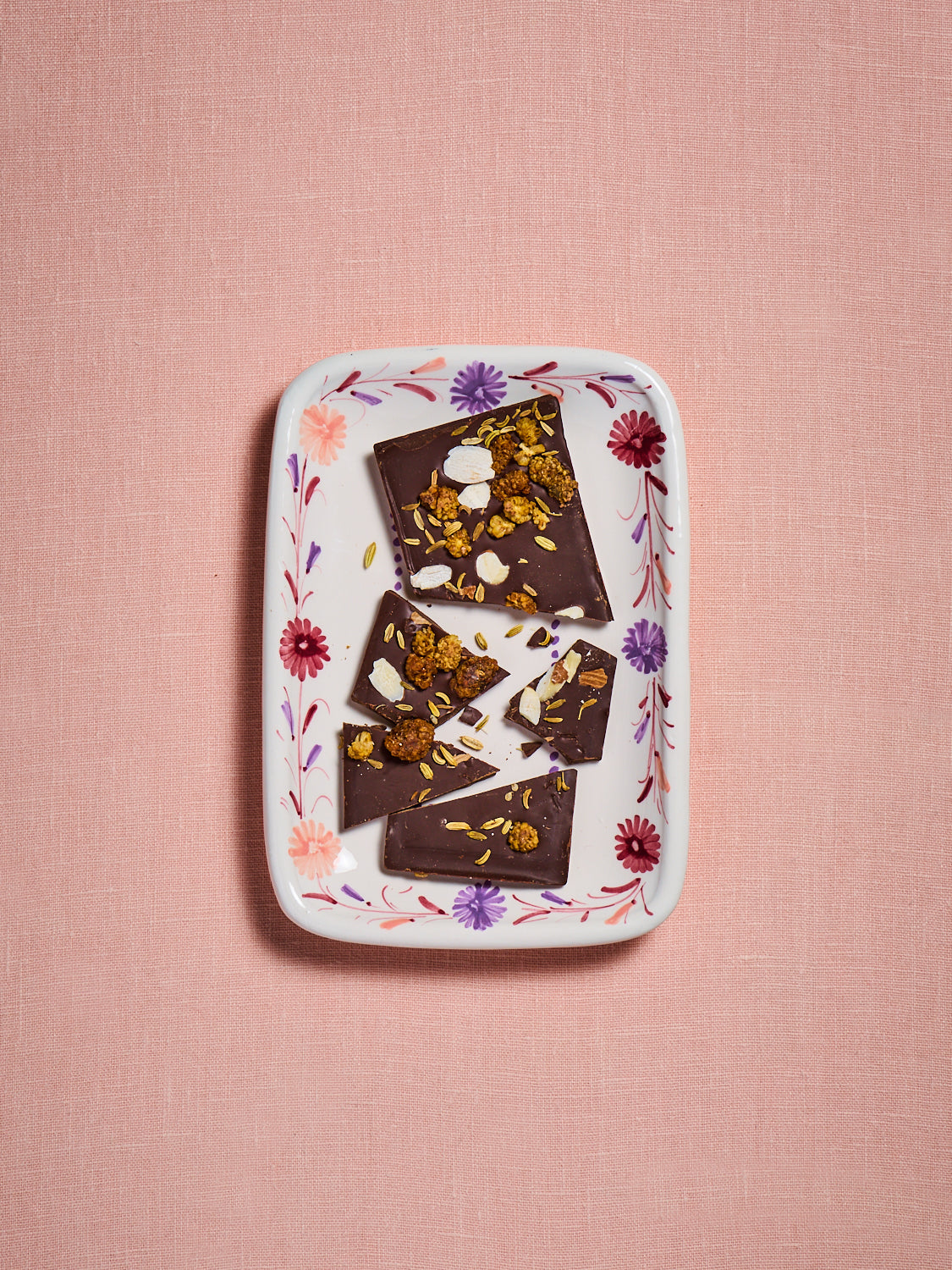 Dark chocolate bar with floral and nut toppings on a hand painted plate with peach, lavender and mulberry flowers against a pink background
