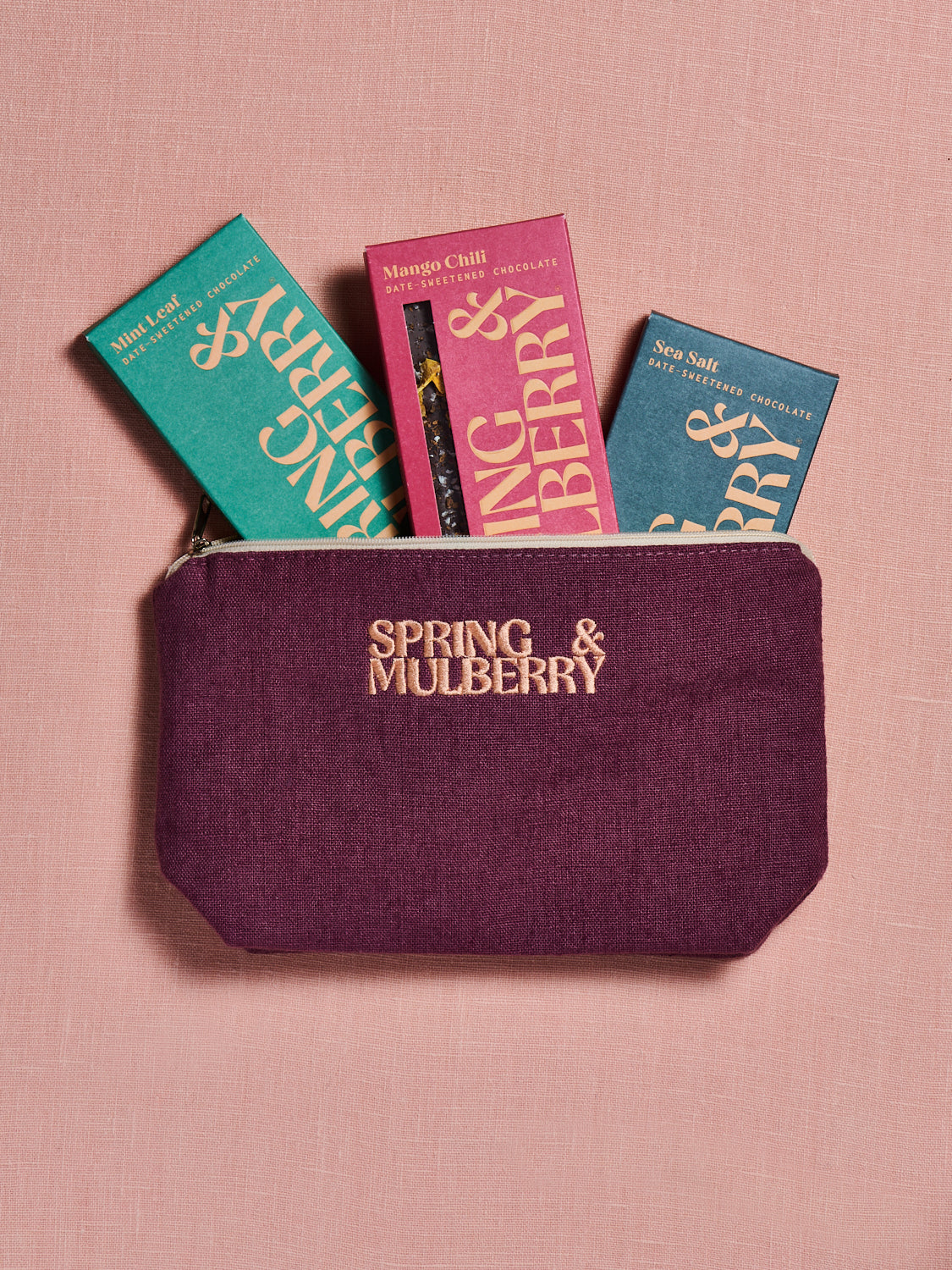 A pouch made to hold chocolate bars shows three different flavors of Spring & Mulberry appearing from the top. The pouch is made of a mulberry linen and embroidered with Spring & Mulberry's logo in peach. The pouch, when sold, does not include the chocolate bars.