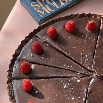Chocolate tart with raspberries and flaky salt on a pink surface, with a Spring & Mulberry pure dark bar in the background. 