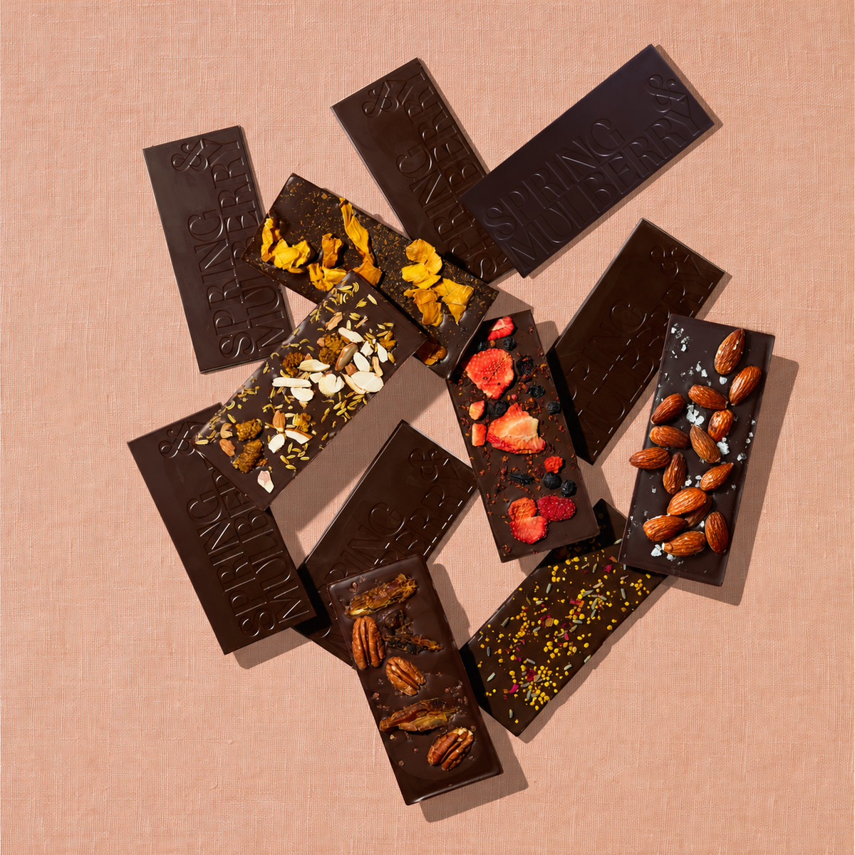 Spring & Mulberry dark chocolate bars arranged in an overlapping flat lay, featuring embossed chocolate and bars topped with almonds, pecans, dried berries, mango, and floral accents against a textured peach background.