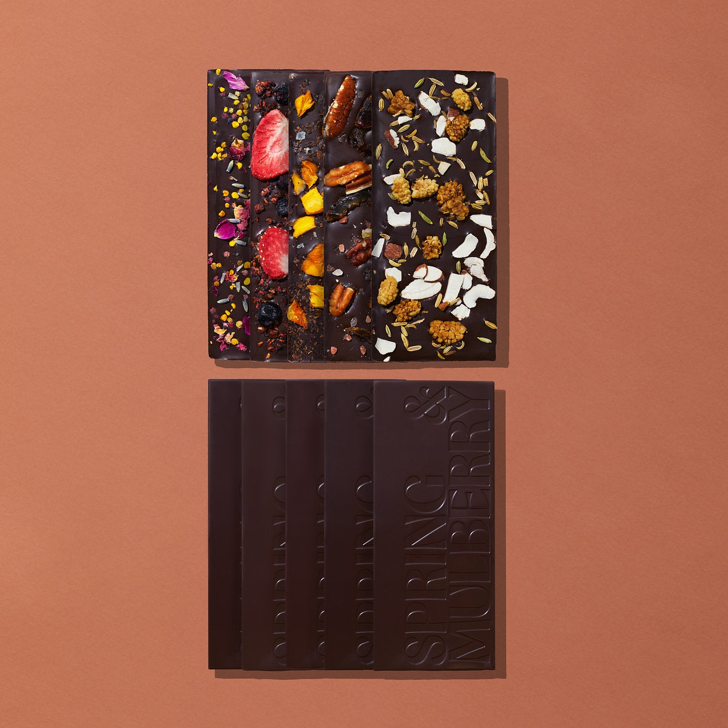 Ten chocolate bars, five topped with fruits, florals, nuts, herbs and spices, and five infused with essential oils, are laid out on a warm brown background.