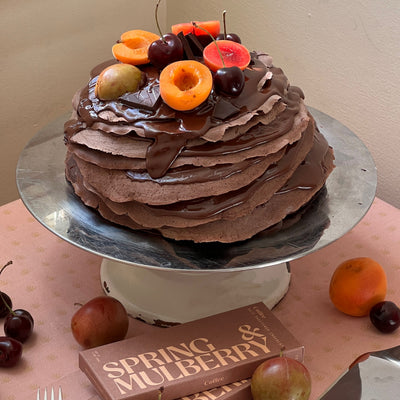 Chocolate cake with fruit on a silver cake stand, surrounded by fruits and a box of Spring Mulberry chocolate.