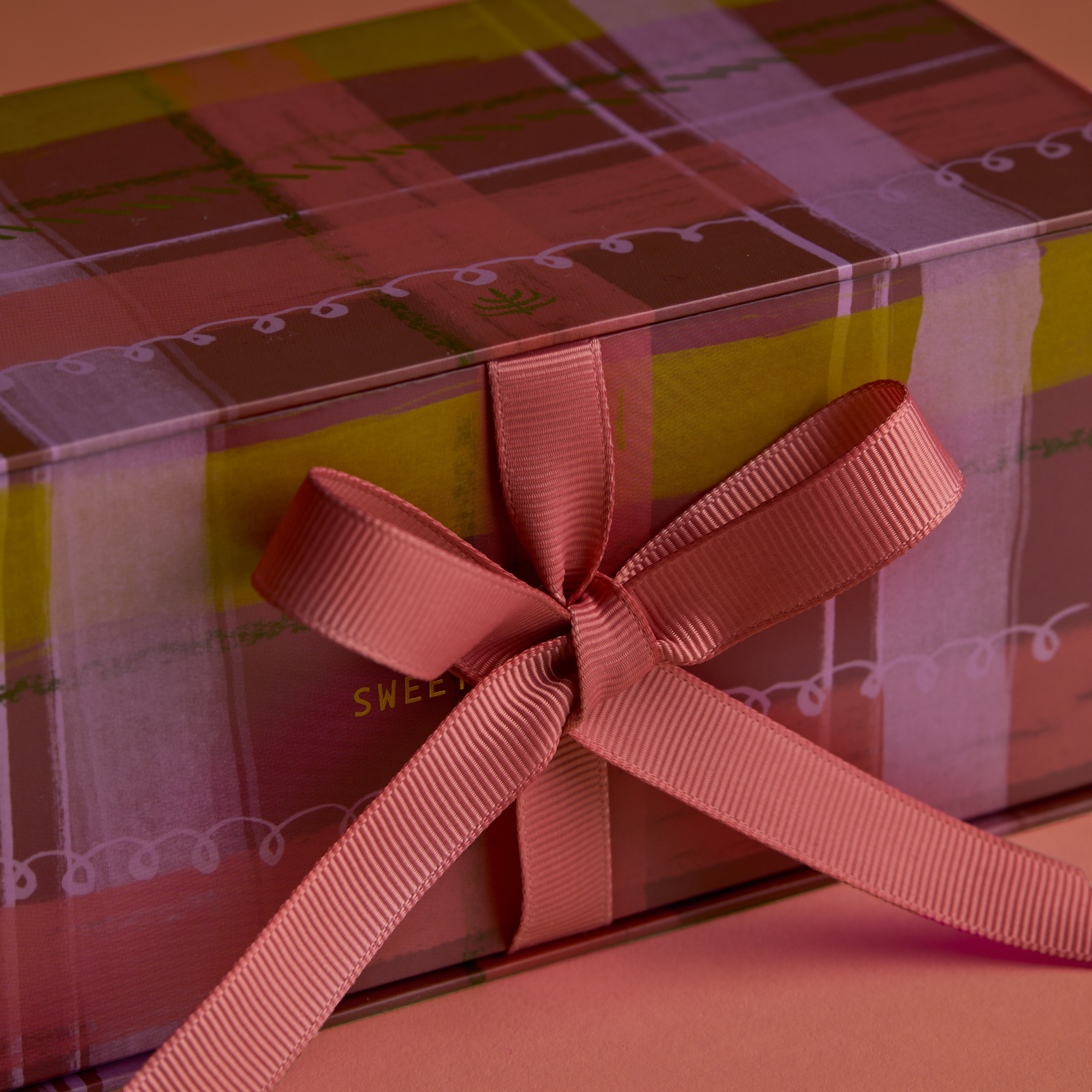 Close up of one gift box featuring a watercolor plaid motif in a rose hue with coordinating pink ribbon that is tied in a bow sitting on a warm brown background. 