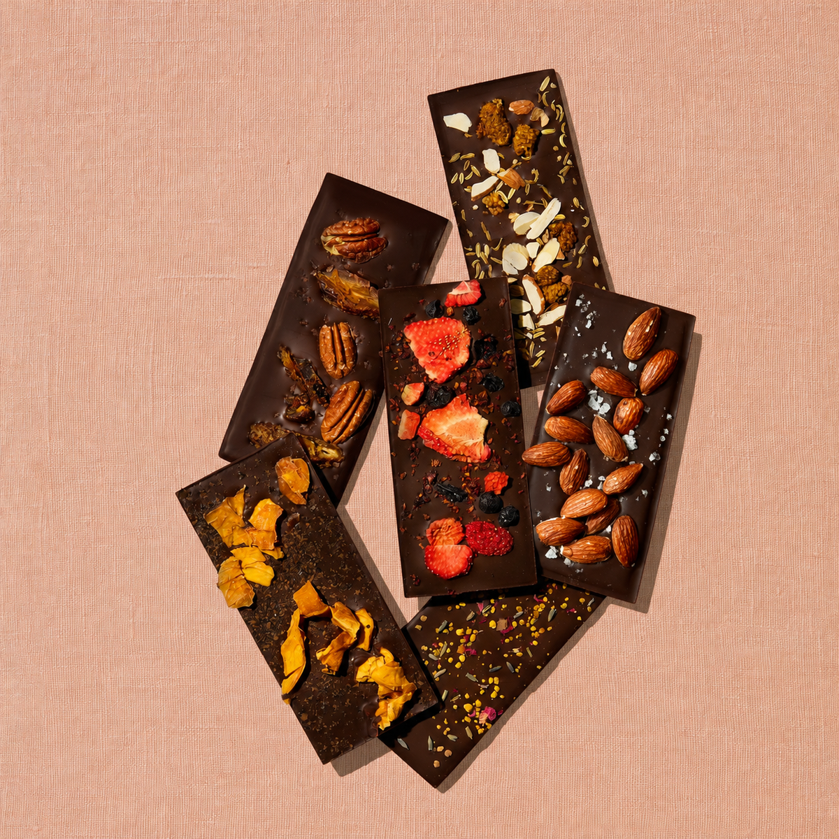Six Spring & Mulberry dark chocolate bars arranged in an overlapping display, topped with dried strawberries, mixed berries, pecans, dates, mango, almonds, flaky sea salt, and floral accents against a soft peach textured background.
