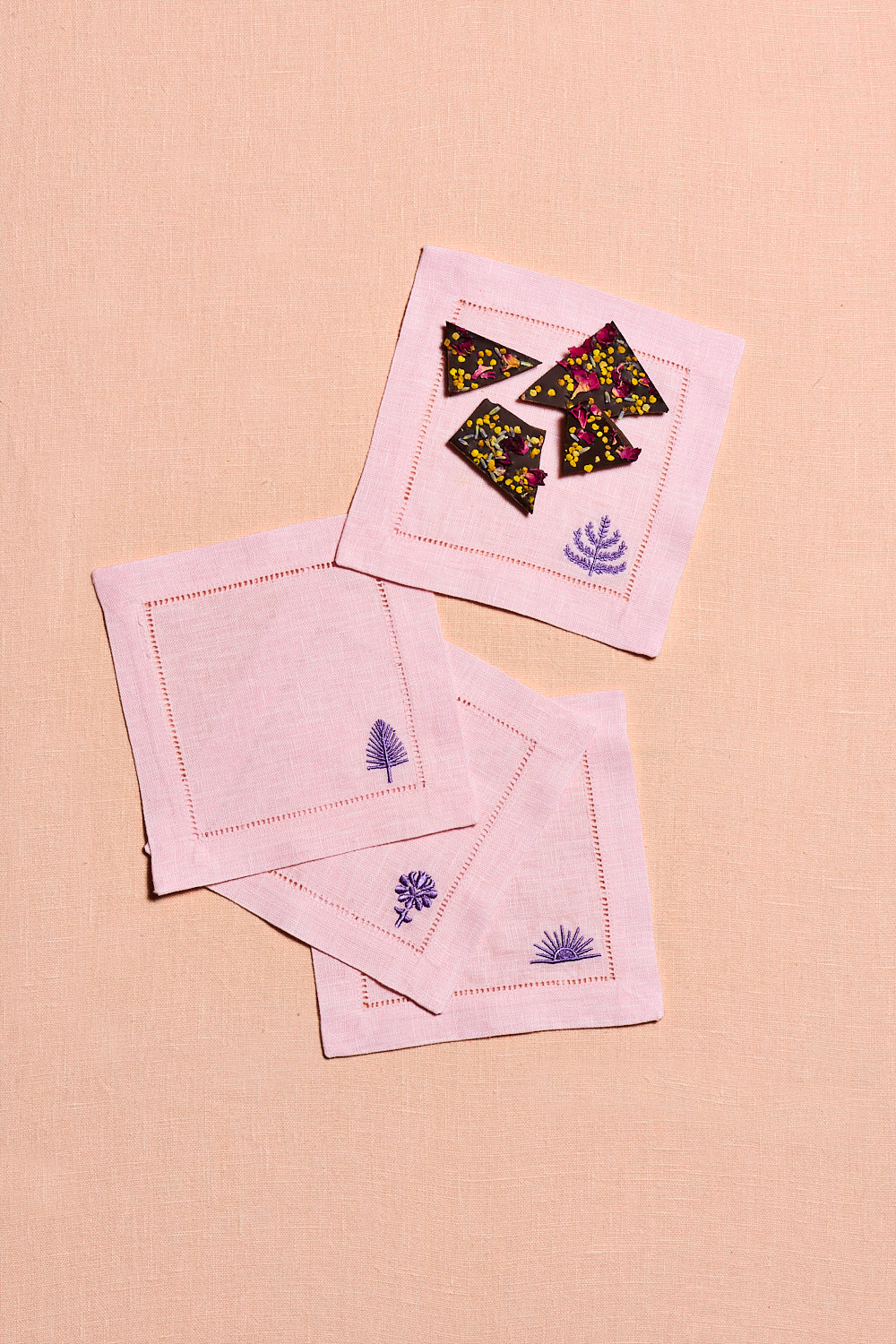 Set of blush pink linen cocktail napkins with embroidered botanical spring & mulberry logos, one napkin has a lavender rose bar and they are sitting on top of a peach background