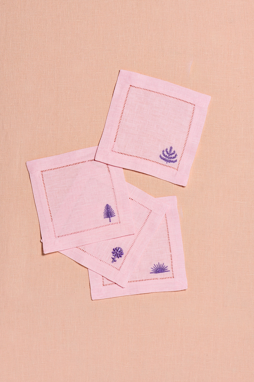 Set of blush pink linen cocktail napkins with embroidered spring & mulberry logos on a soft peach backdrop.