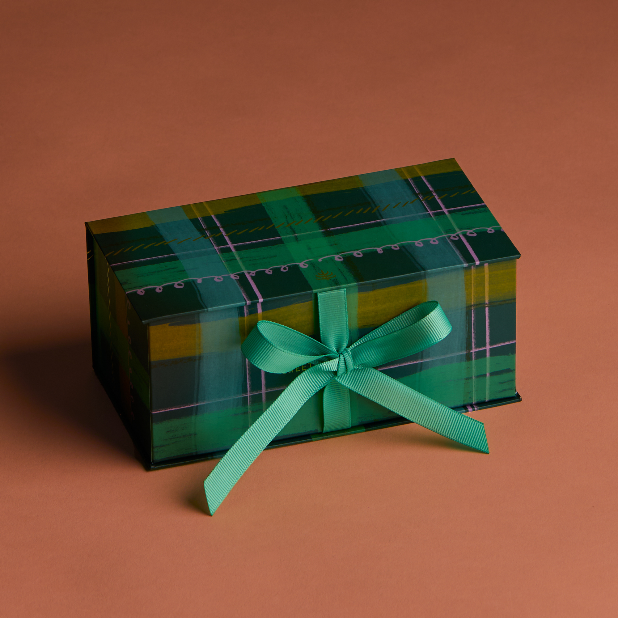 One gift box featuring a watercolor plaid motif in a blue hue with coordinating green ribbon that is tied in a bow sitting on a warm brown background. 