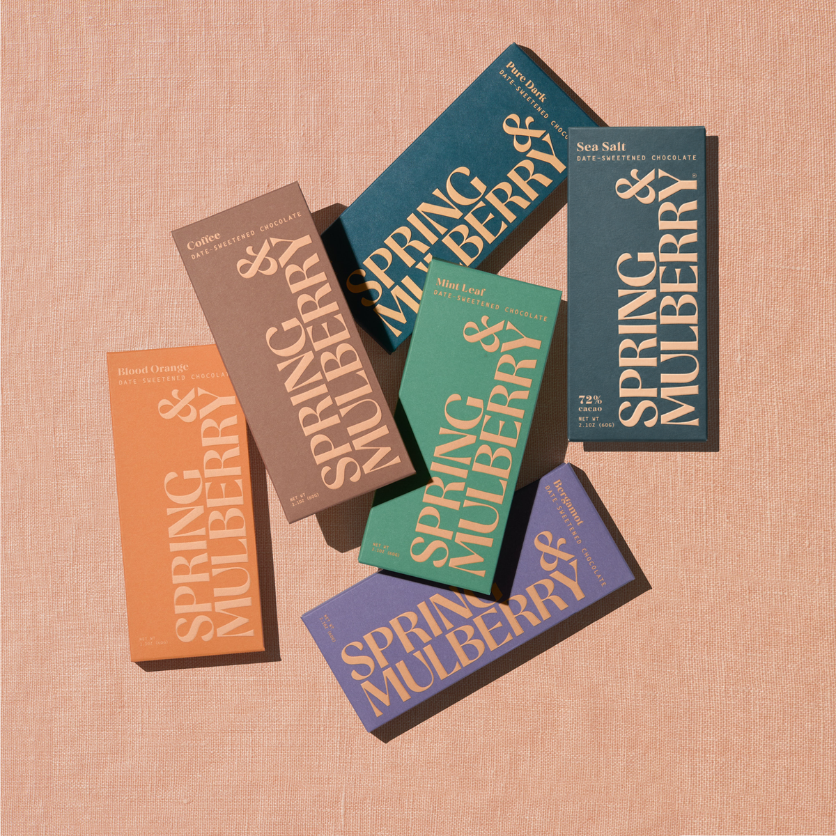 Five artisanal chocolate bars in their Spring & Mulberry boxes are laid out on a surface, each with different color packaging representing various flavors.