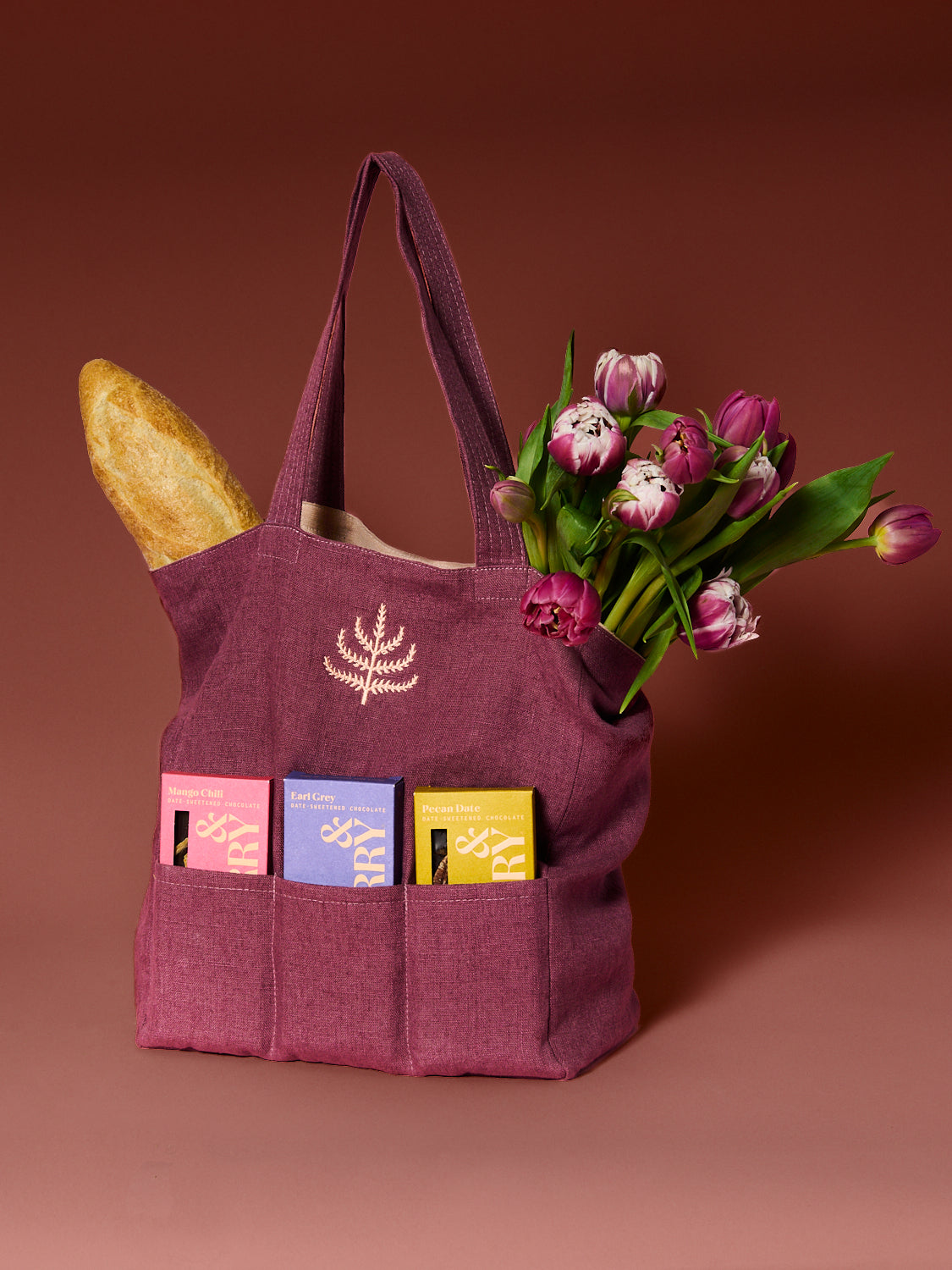 Mulberry tote bag designed with three chocolate compartments filled with chocolate, bread, and flowers on a rose hued background.