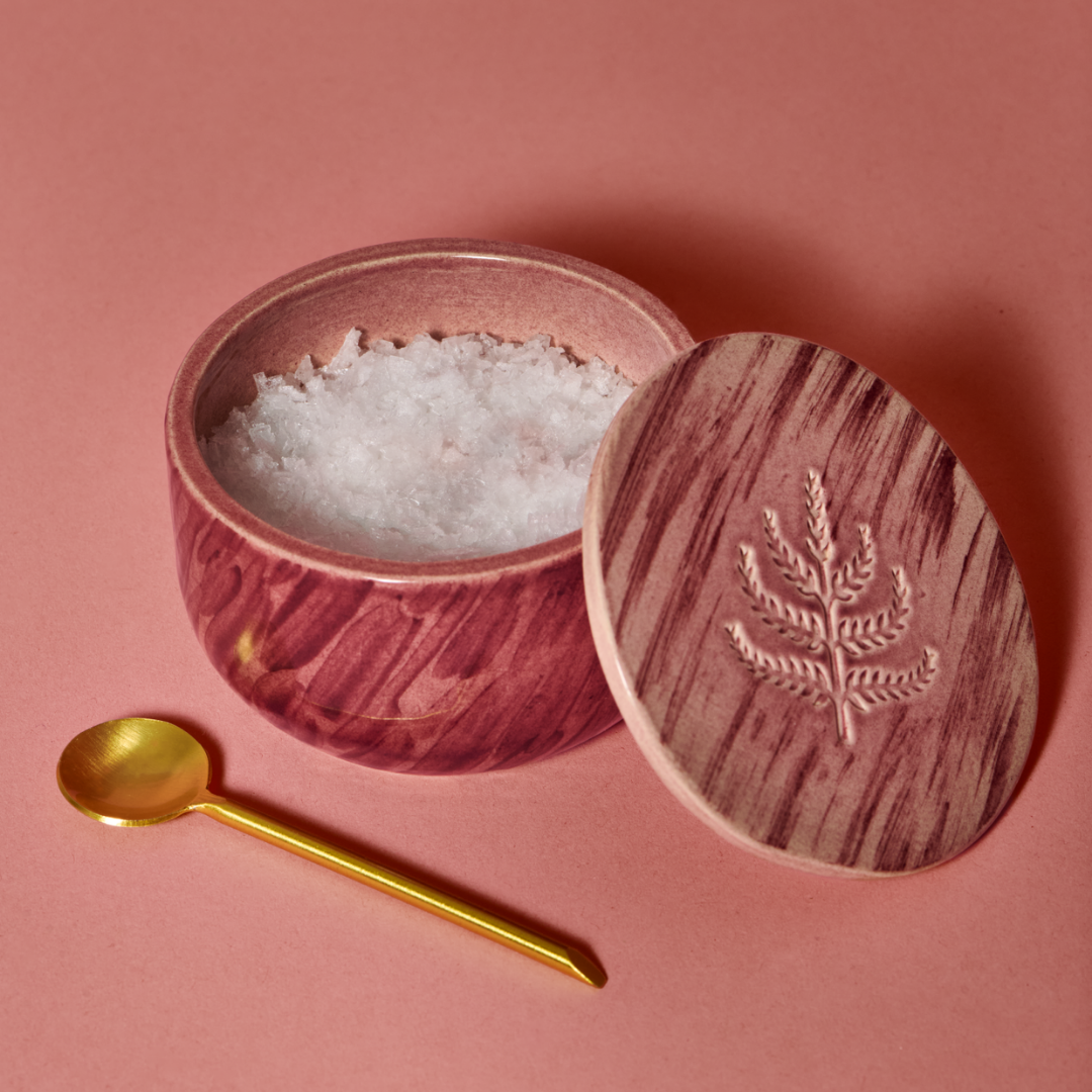 Flake salt sits in a handmade salt cellar with imprinted mulberry tree on the lid. An accompanying gold spoon rests to the side.