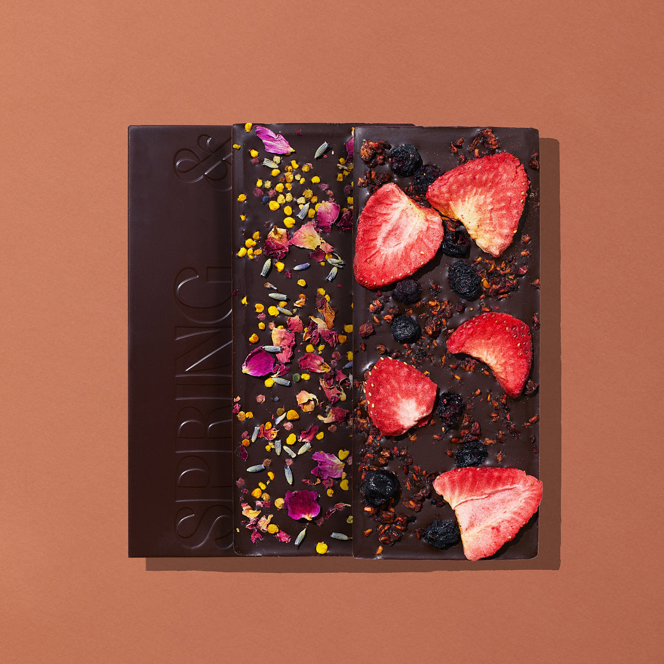 A set of three artisanal chocolate bars layed out flat on a warm brown background, two of which are displaying their toppings. The bars include Pure Dark (no toppings), Lavender Rose, and Mixed Berry.