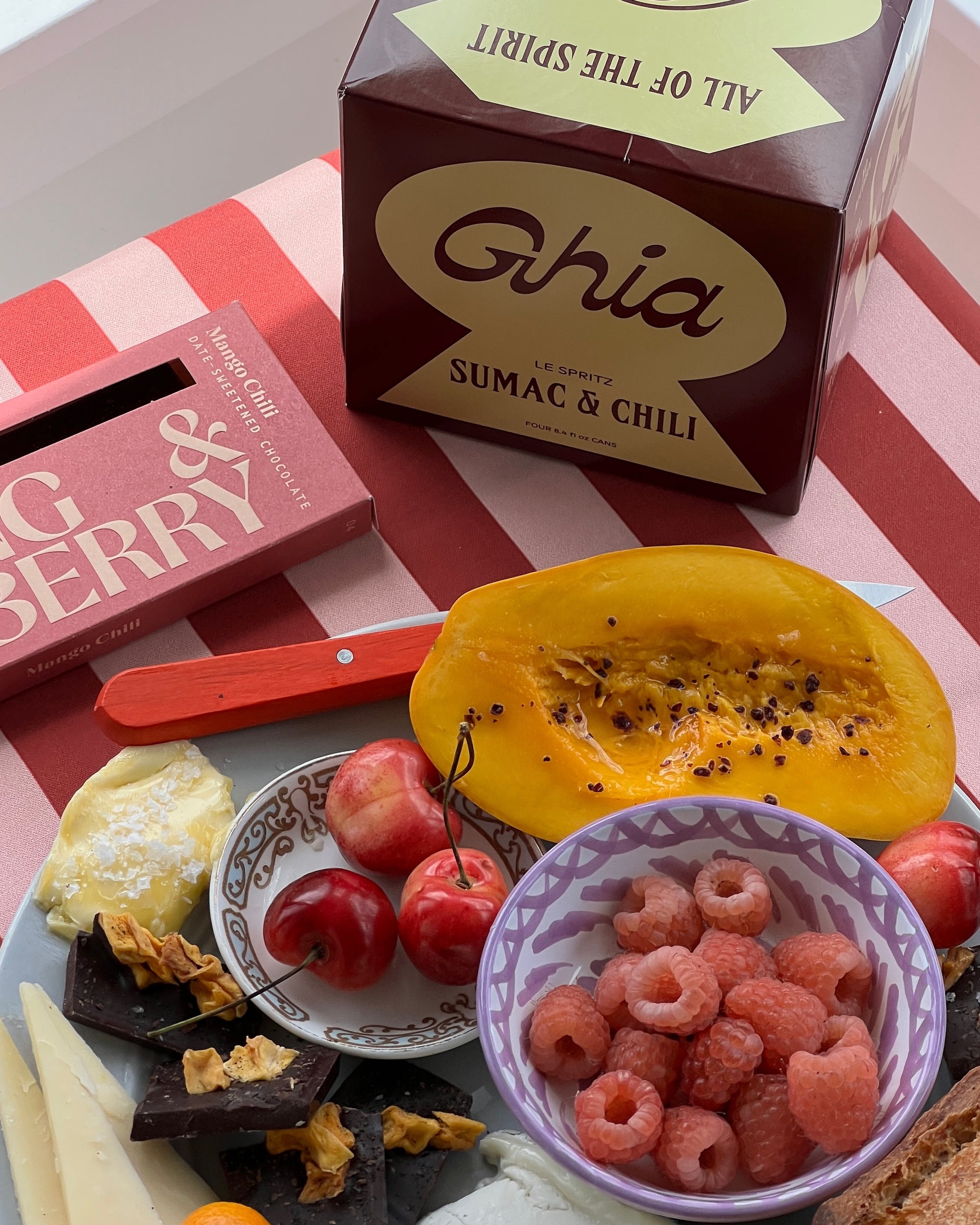 Ghia sumac and chili  4 pack, next to a mango chili bar and a platter of fruit, cheese and chocolate on top of a striped tablecloth