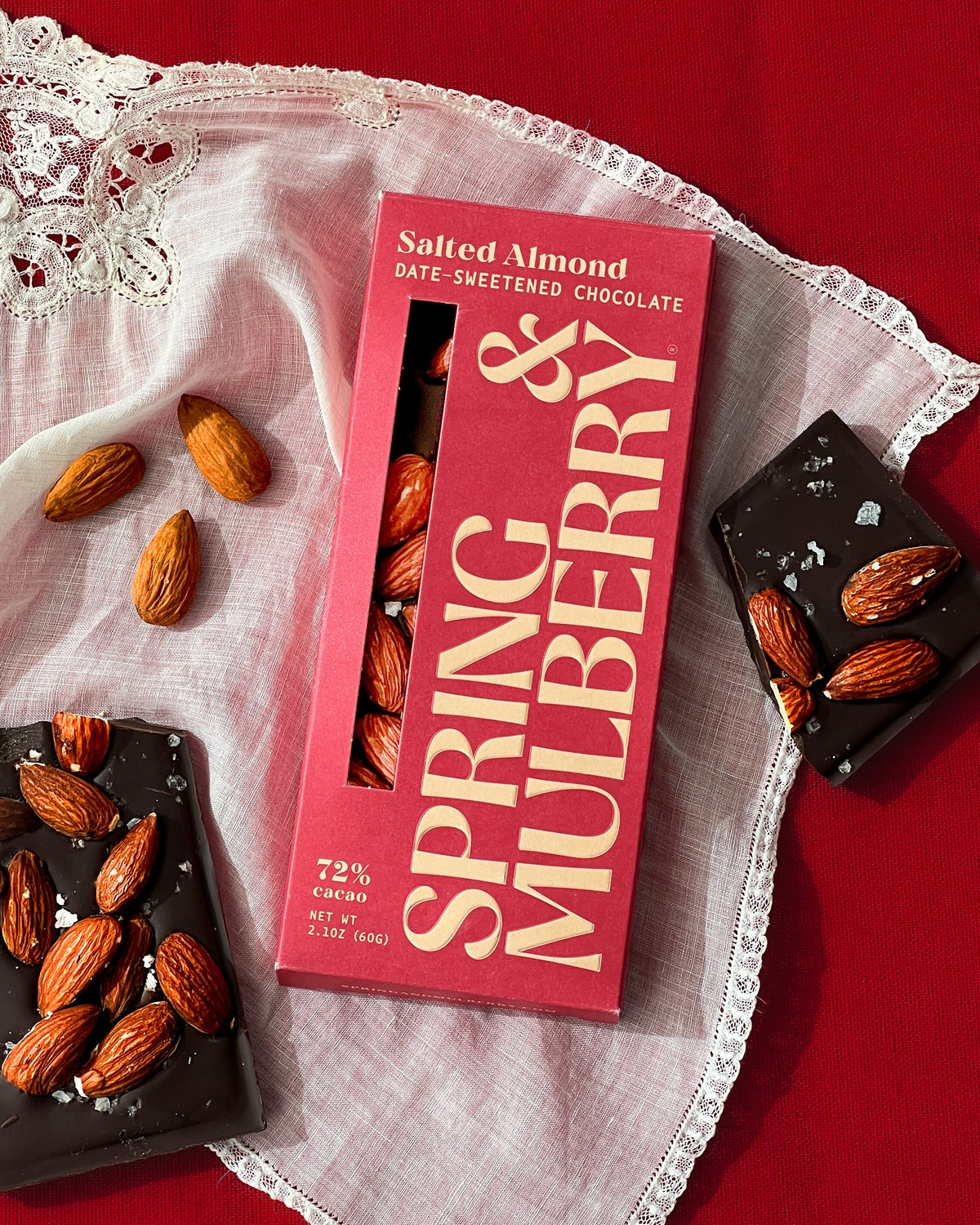 A red Salted Almond bar box sits upon a red background and white vintage hankerchief. Alongisde the bar are broken pieces of Salted Almond chocolate and a few almonds. 