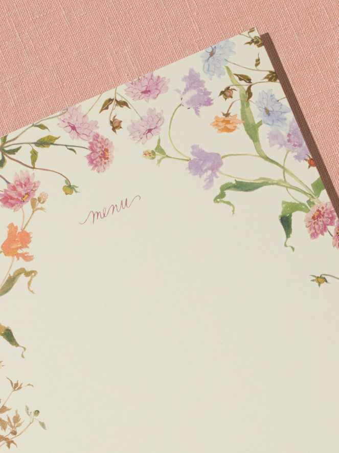A close-up of a menu card sits on a peach, linen backdrop. The menu card features a border of handpainted florals by Raleigh-based artist Inslee Fariss. The flowers are inspired by the sunset-colored packaging of Spring & Mulberry chocolate bars.
