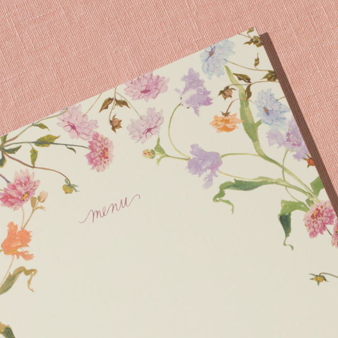 A close-up of a menu card sits on a peach, linen backdrop. The menu card features a border of handpainted florals by Raleigh-based artist Inslee Fariss. The flowers are inspired by the sunset-colored packaging of Spring & Mulberry chocolate bars.