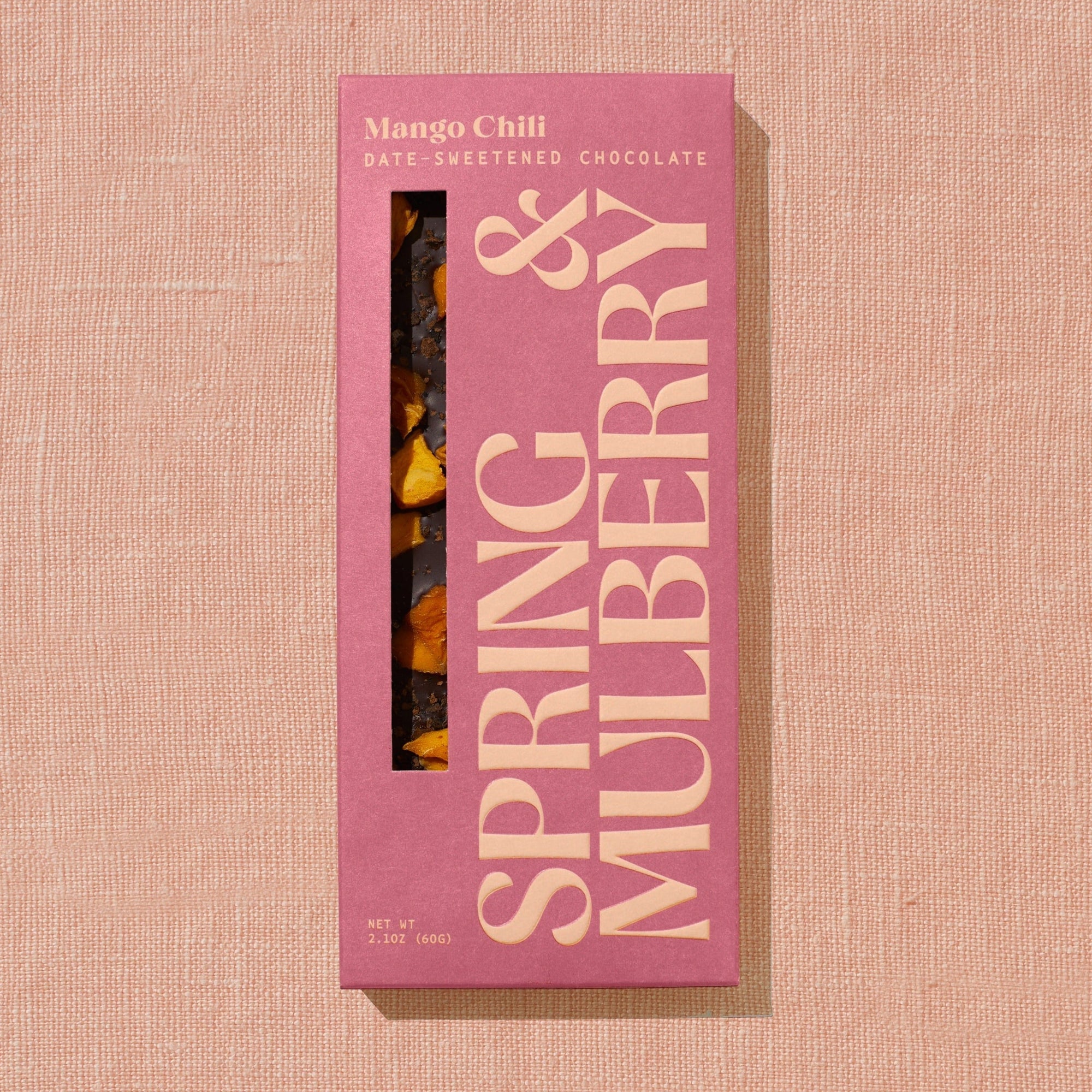 A red/orange Spring & Mulberry box labled Mango Chili date-sweetened chocolate is laid on a warm brown background. 