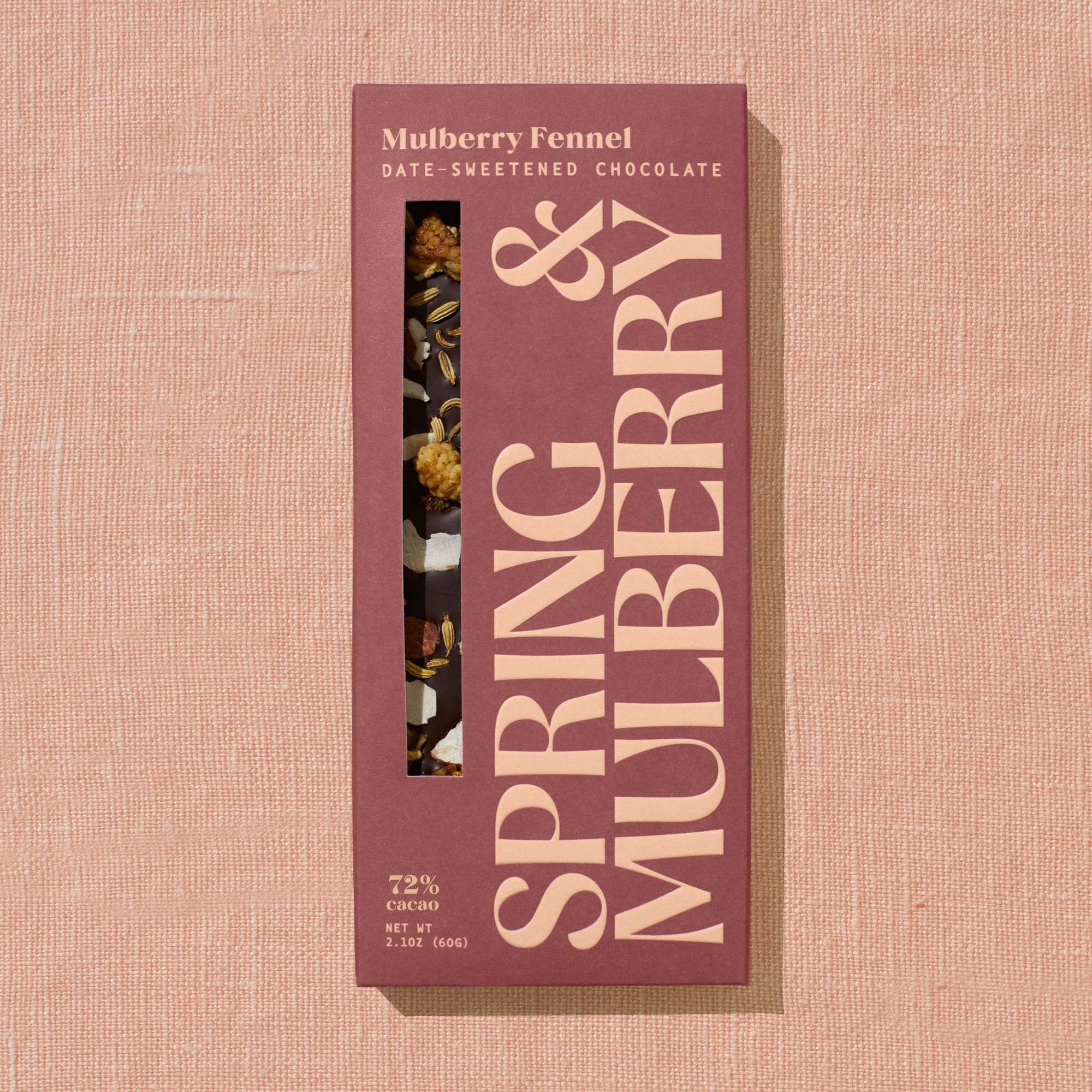 A brick red Spring & Mulberry box labled Mulberry Fennel date-sweetened chocolate is laid on a warm brown background. 