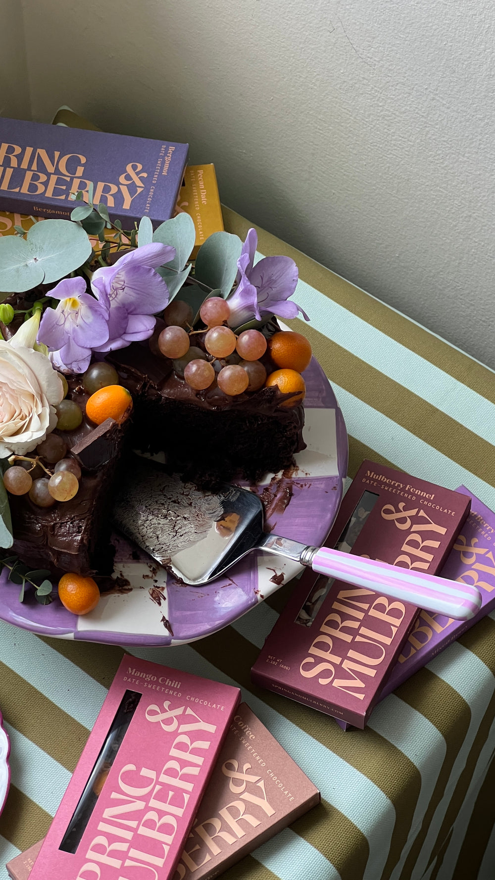 An Olive Oil Chocolate cake adorned with fresh fruit and florals, sits elegantly with a single slice removed—revealing its rich interior. It’s surrounded by Spring & Mulberry chocolate bars, artfully arranged on a striped tablecloth.