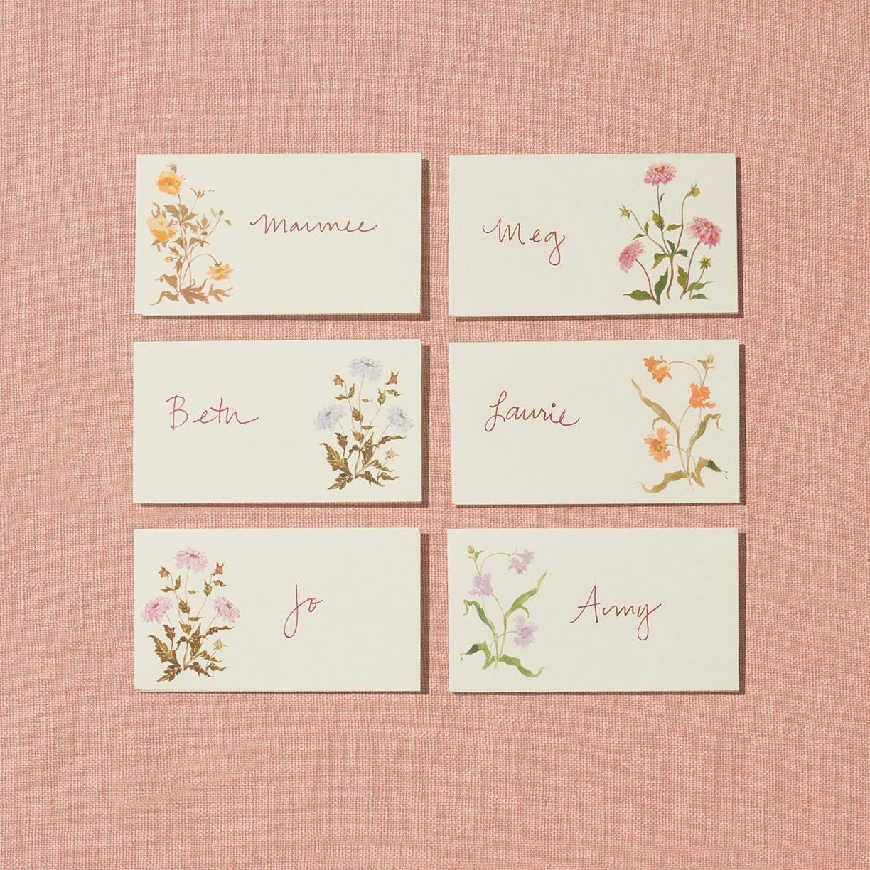 A set of six creamy white placecards with hand-painted pink, purple, and orange floral designs on one side of the card with room for personalized names on the other; these are displayed on a pink background.