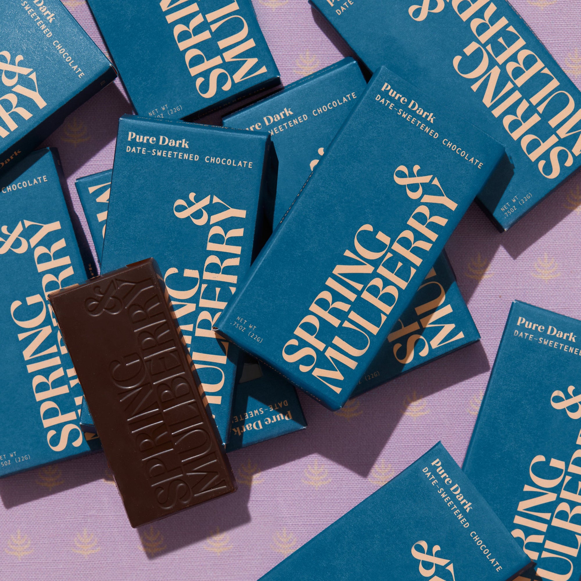 A dozen Pure Dark Mini chocolate boxes sporadically laid out on a pink background with cream mulberry logos patterned out. One bar is out of its packaging and displaying its stamped logo. 