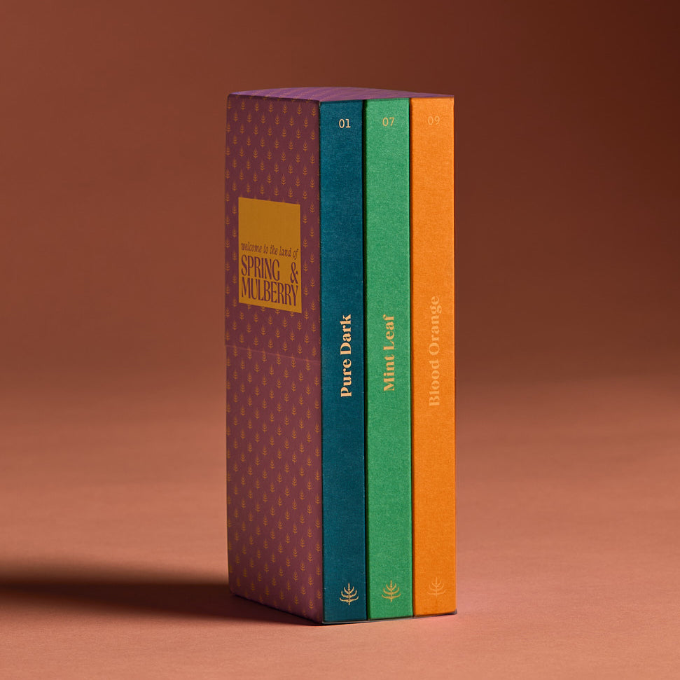 A set of three artisanal chocolate bars in a vertical band, with individual bars labeled Pure Dark, Mint Leaf, and Blood Orange, indicating different flavors. The band is lavender with a peach mulberry motif and Spring & Mulberry logo.