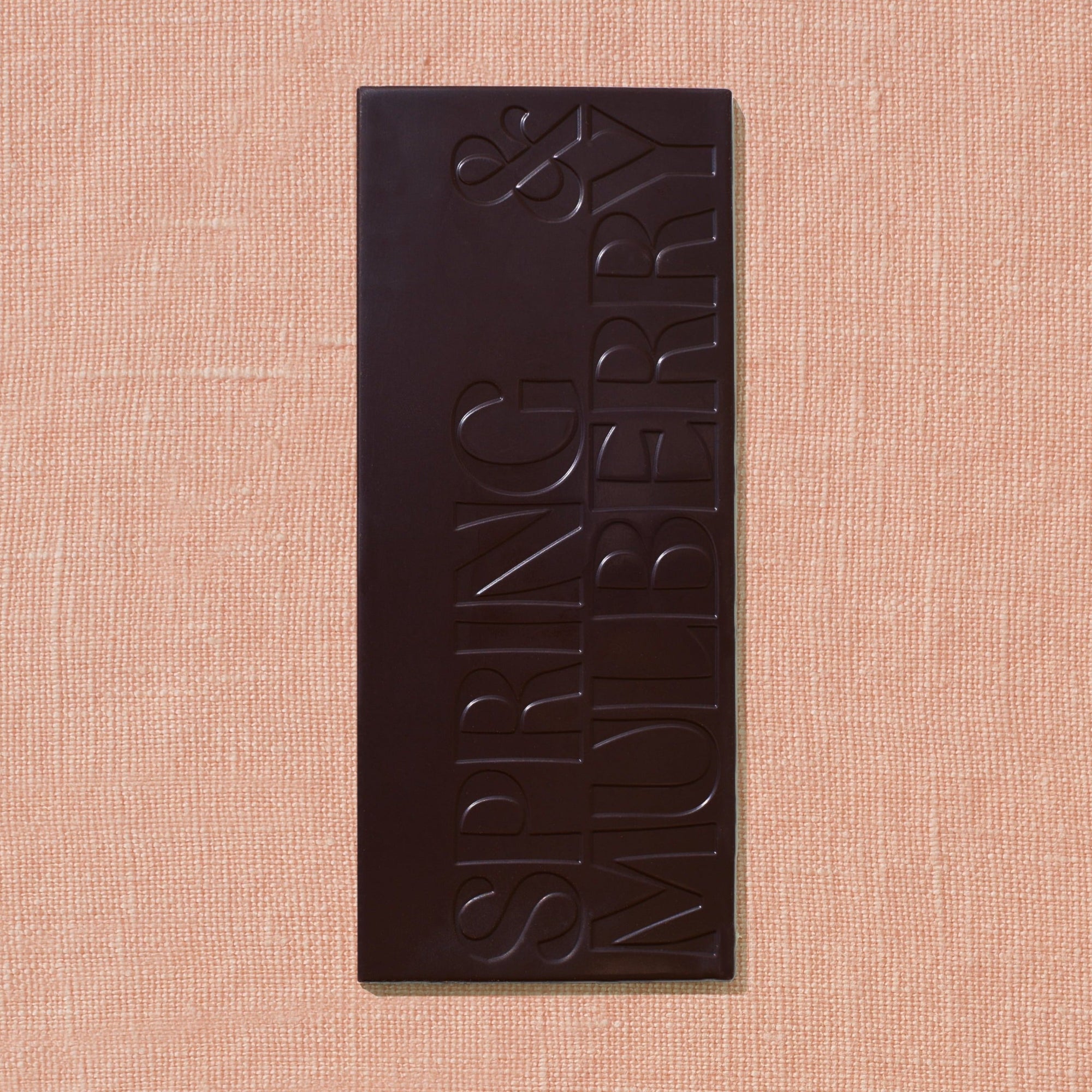 One chocolate bar that is stamped with Spring & Mulberry logo is laid out on a warm brown background.