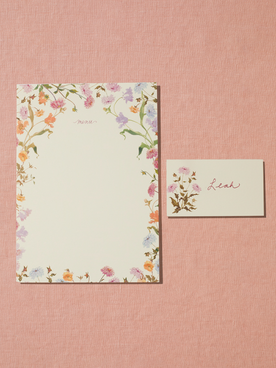 A menu and placecard sit on a linen peach backdrop. The menu card on the left features a border of handpainted florals on a creamy white card. The placecard on the right features one pink flower and a handwritten name.