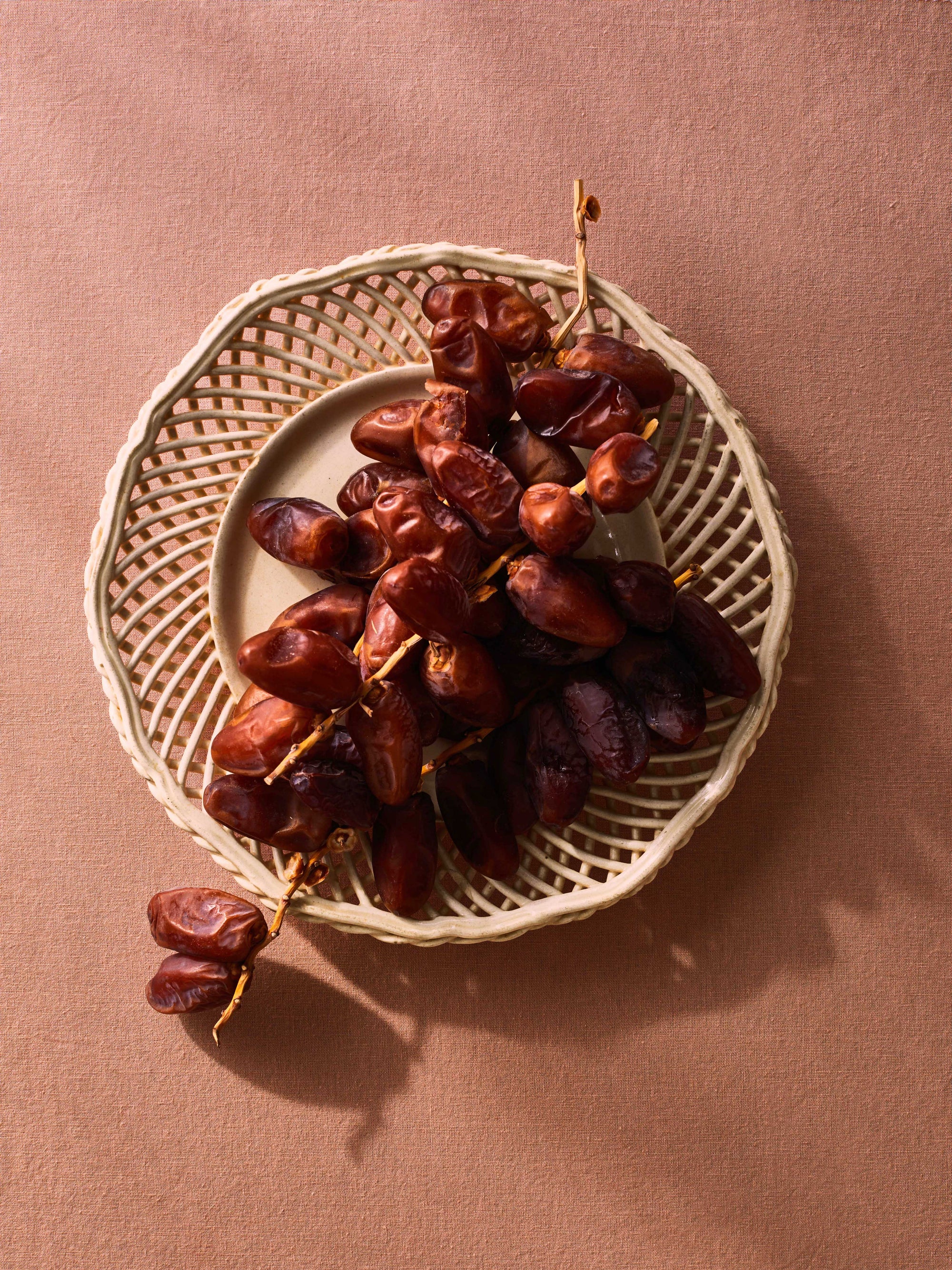 Shot with an overhead view, a peach linen tablecloth is the backdrop a white lattice porcelain plate filled with an overflowing bunch of dates, still on the branch. Warm light comes from the upper left, casting a moody shadow on the bottom right of the image, indicating evening.