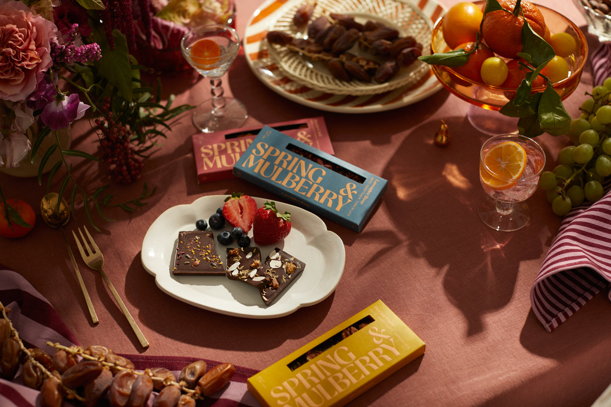 A berry-topped chocolate bar on a purple Spring & Mulberry box, surrounded by flowers, fruit and drinks on a pink table