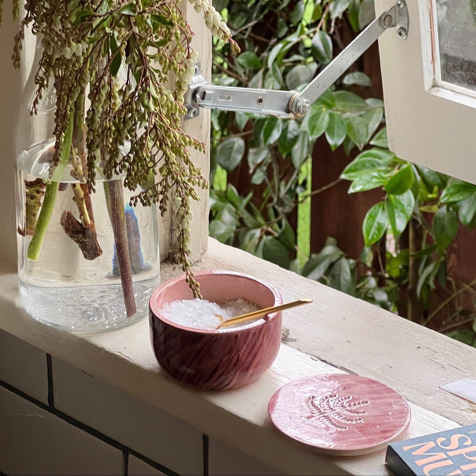Pink ceramic salt cellar filled with flake salt and mini gold spoon sits next to an open window and vase of flowers. 