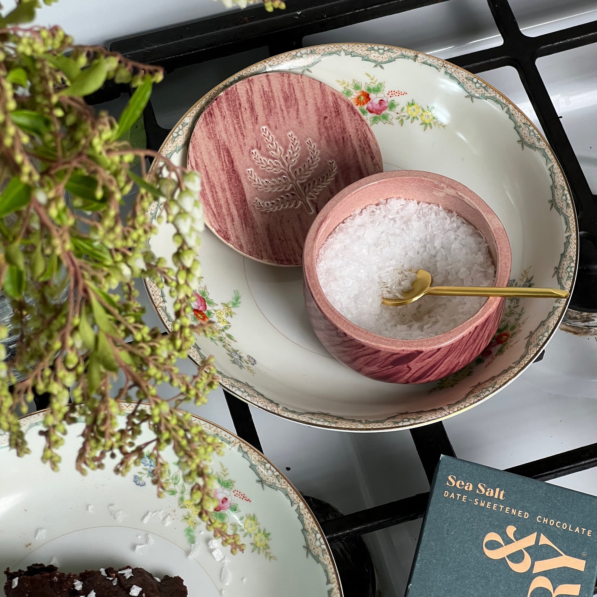 Pink ceramic salt cellar filled with flake salt and mini gold spoon sit on a vintage plate next to a Spring & Mulberry Sea Salt date-sweetened chocolate bar.