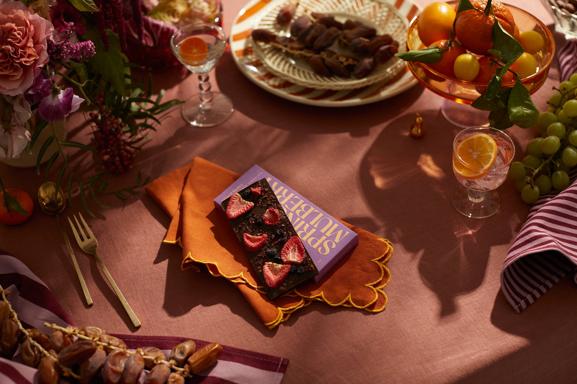 Tablescape with florals and fruits on a pink tablecloth. In the center, a mixed berry bar on top of a scalloped orange napkin. 