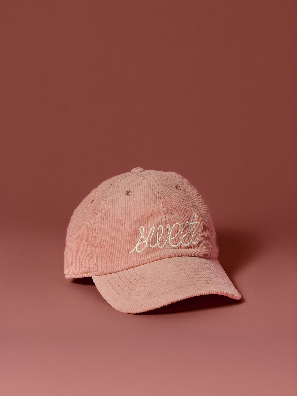 Peach corduroy baseball cap embroidered with the word “sweet” in cream script, photographed against a soft rose-colored background