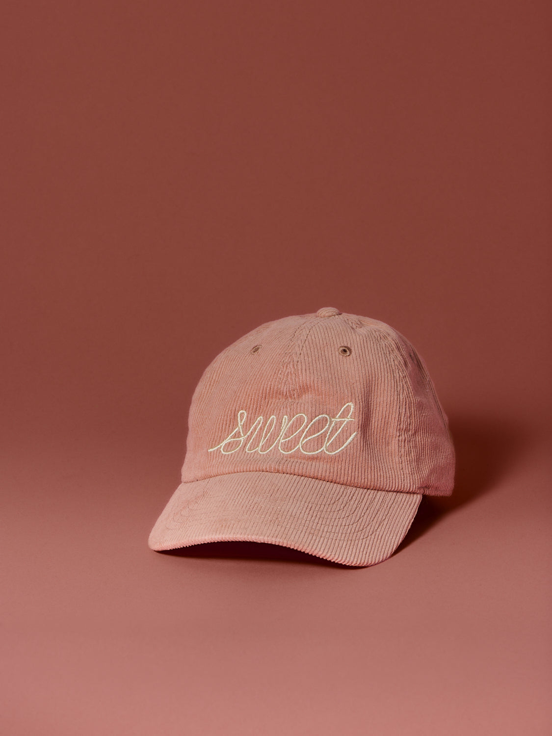 Blush pink corduroy baseball cap embroidered with the word “sweet” in cream script, photographed against a soft rose-colored background