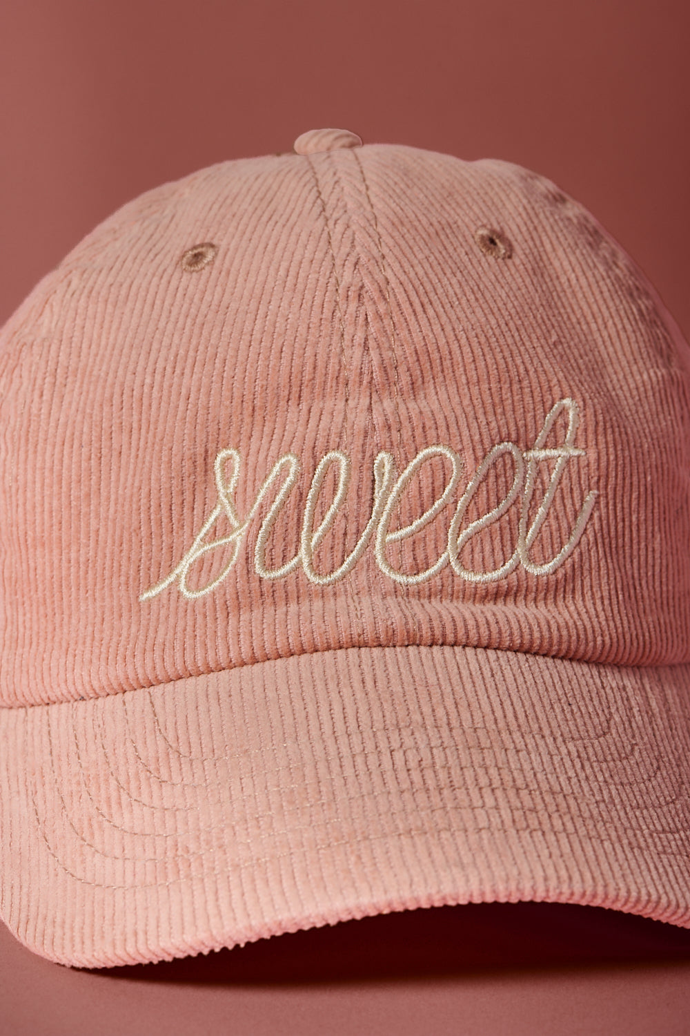 Blush pink corduroy baseball cap embroidered with the word “sweet” in cream script, photographed against a soft rose-colored background
