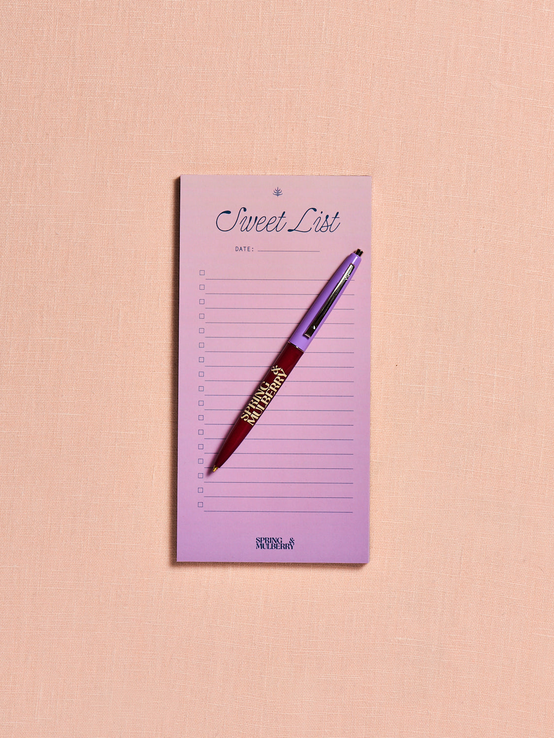 Spring & Mulberry "Sweet List" notepad in soft lavender with a matching branded pen, featuring a checklist layout for notes, errands, and grocery lists on a blush pink background.