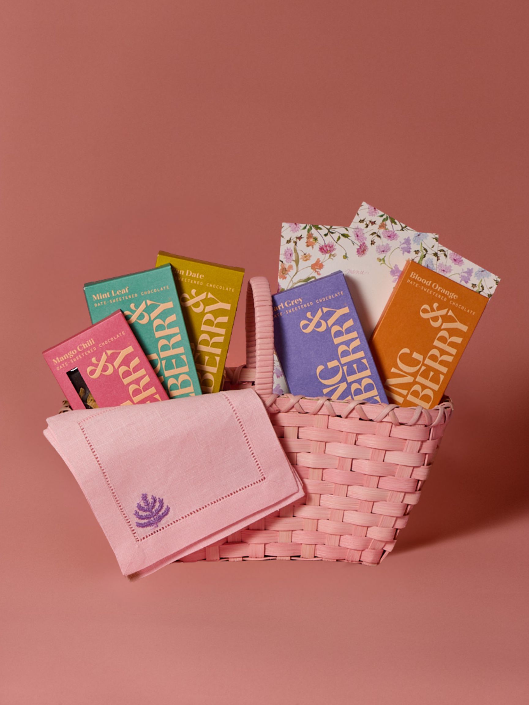 Handmade pink woven rattan handbag on a matching pink background. Designed for Spring & Mulberry by Underwater Weaving. Filled with five date-sweetened chocolate bars, floral menu cards, and linen chocolate napkins.