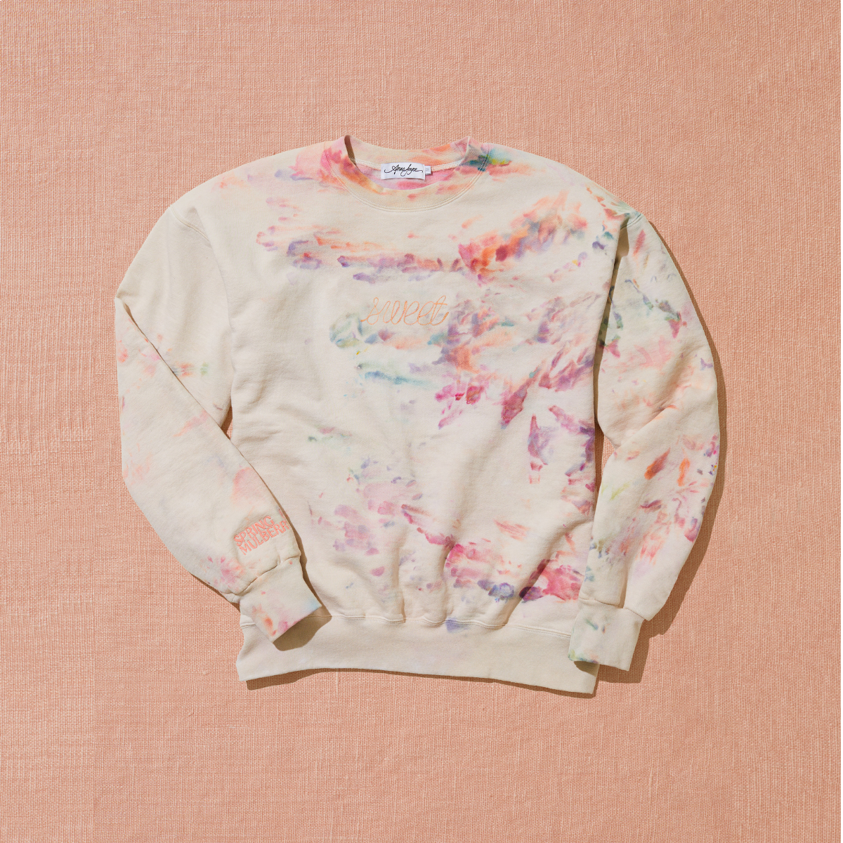 A cream based sweetshirt with 'Sweet' embroidered in light pink across the chest and also hand dyed in the colors of pink, orange, purple, and light blue by using a unique ice technique is laid out on a warm brown background.  