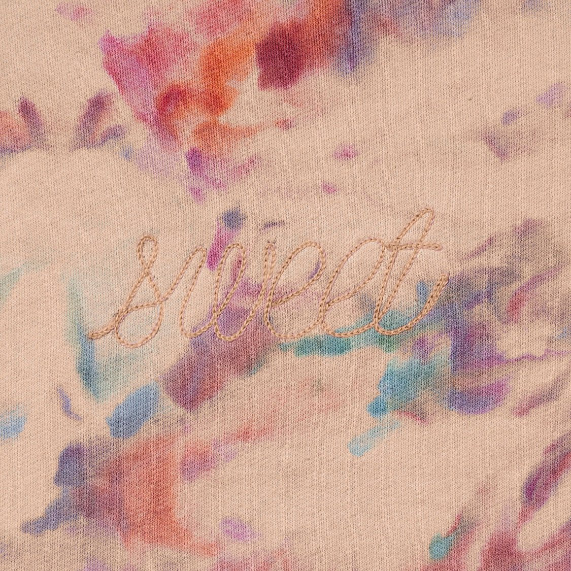 A close up shot of the peach based sweetshirt with 'Sweet' embroidered in light pink across the chest and also hand dyed in the colors of pink, orange, purple, and light blue by using a unique ice technique.  