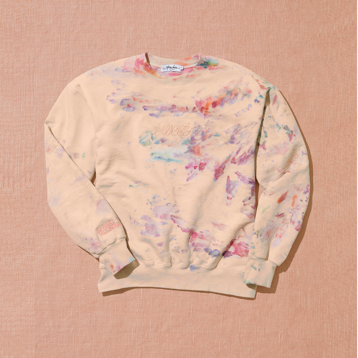 A peach based sweetshirt with 'Sweet' embroidered in light pink across the chest and also hand dyed in the colors of pink, orange, purple, and light blue by using a unique ice technique is laid out on a warm brown background.  
