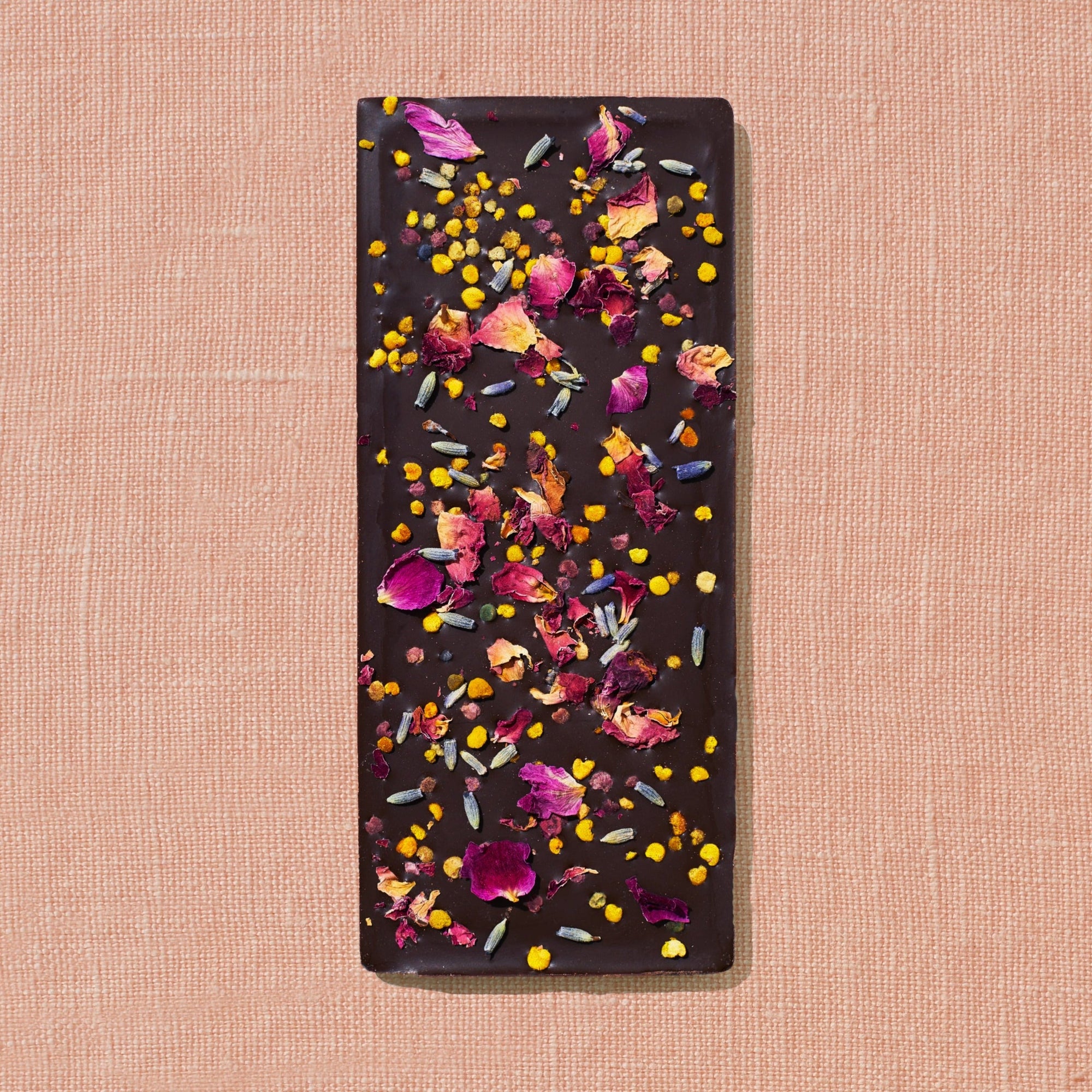 One chocolate bar that is topped with lavender, rose petals, and bee pollen is laid out on a warm brown background.