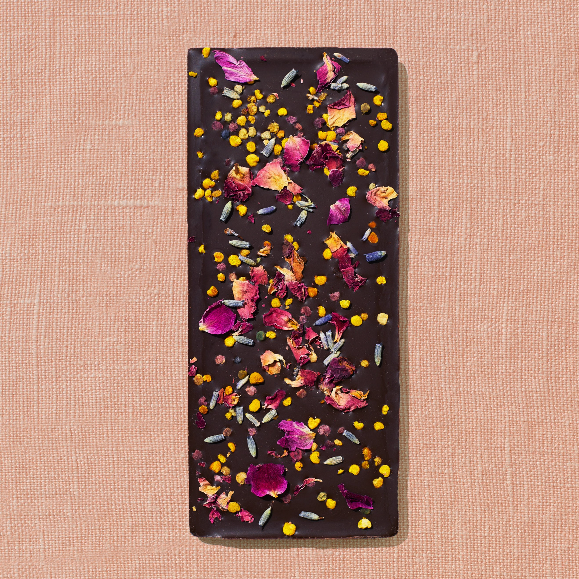 One chocolate bar that is topped with lavender, rose petals, and bee pollen is laid out on a warm brown background.