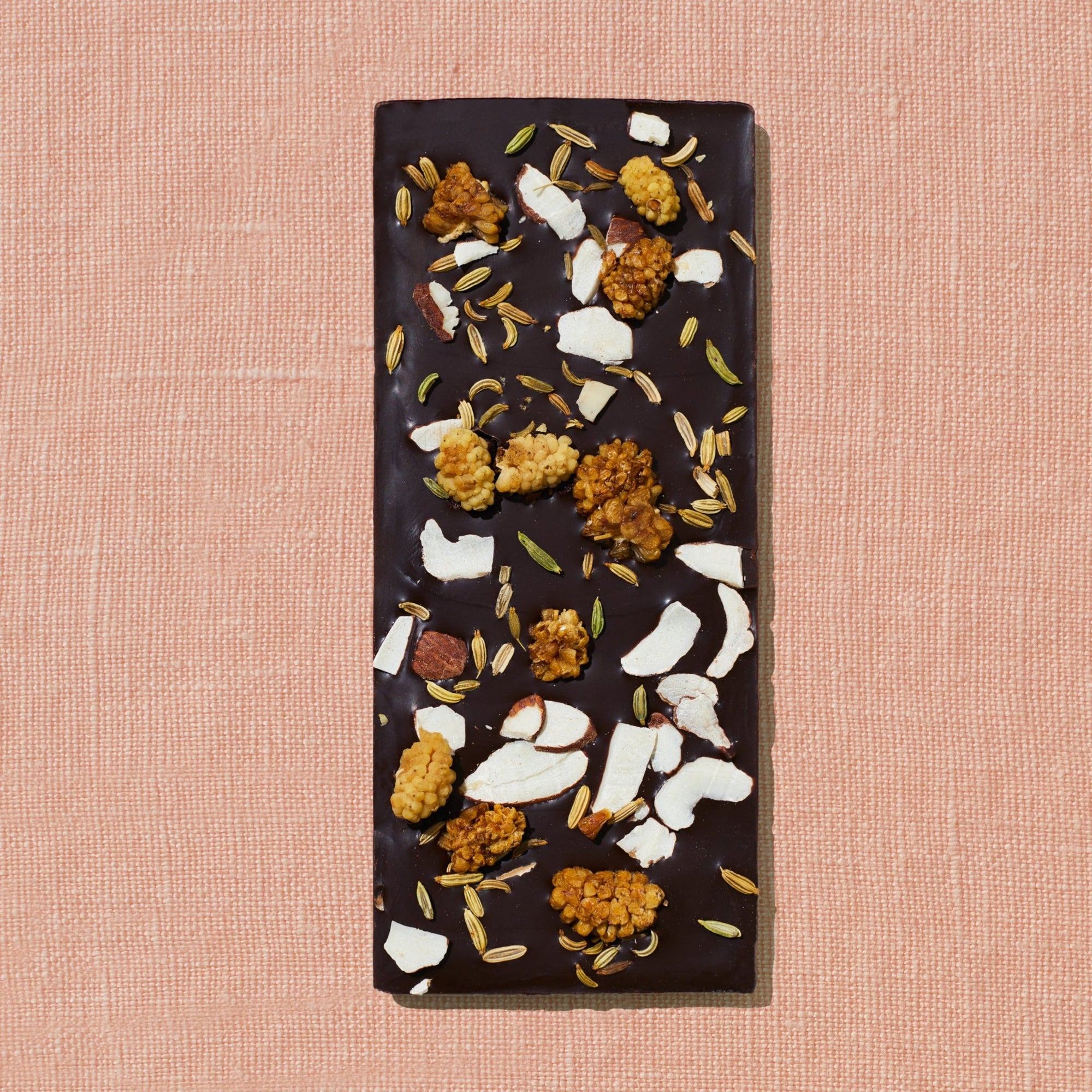 One chocolate bar that is topped with mulberries, almond flakes, and fennel is laid out on a warm brown background.