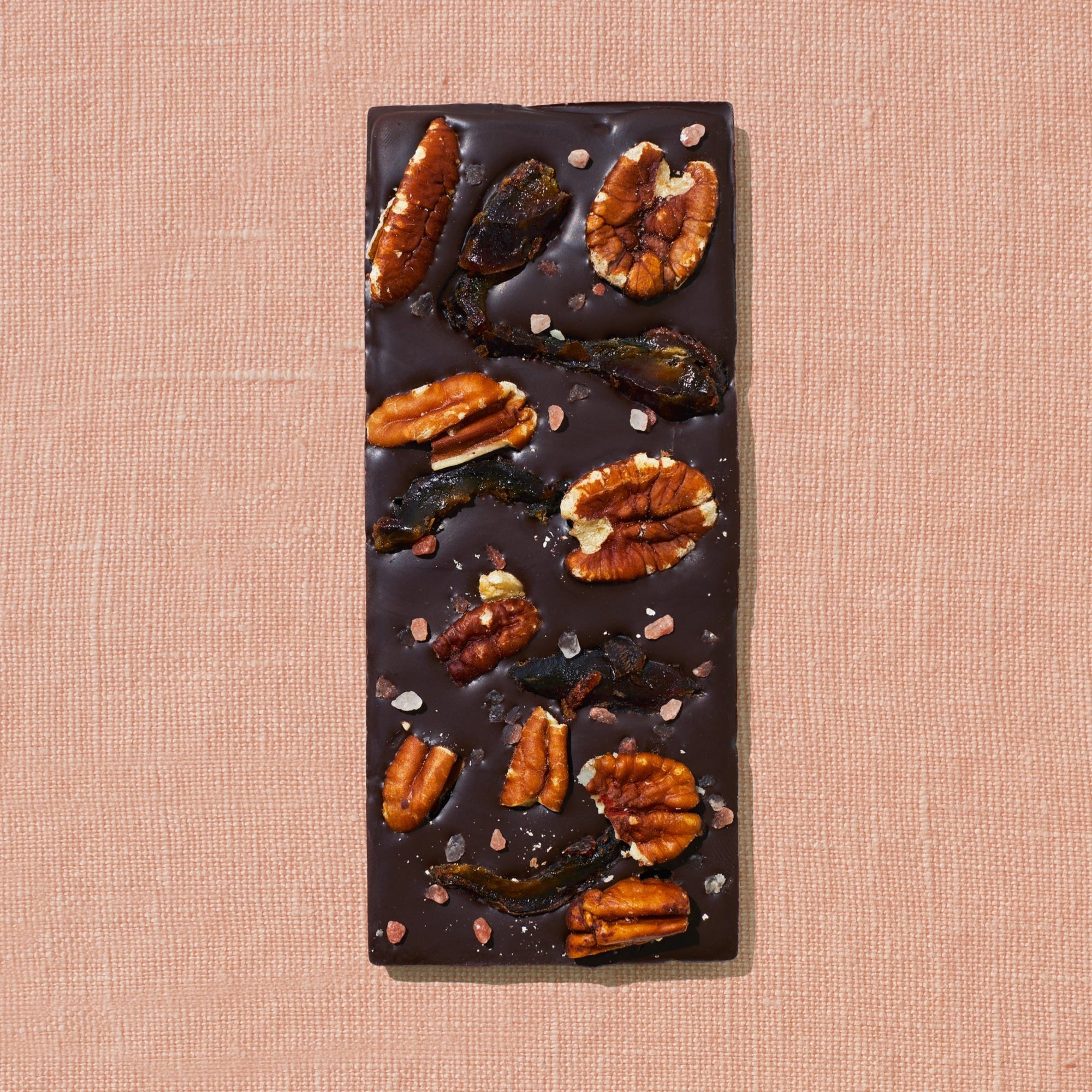 One chocolate bar that is topped with dates, pecans, and rock salt is laid out on a warm brown background.