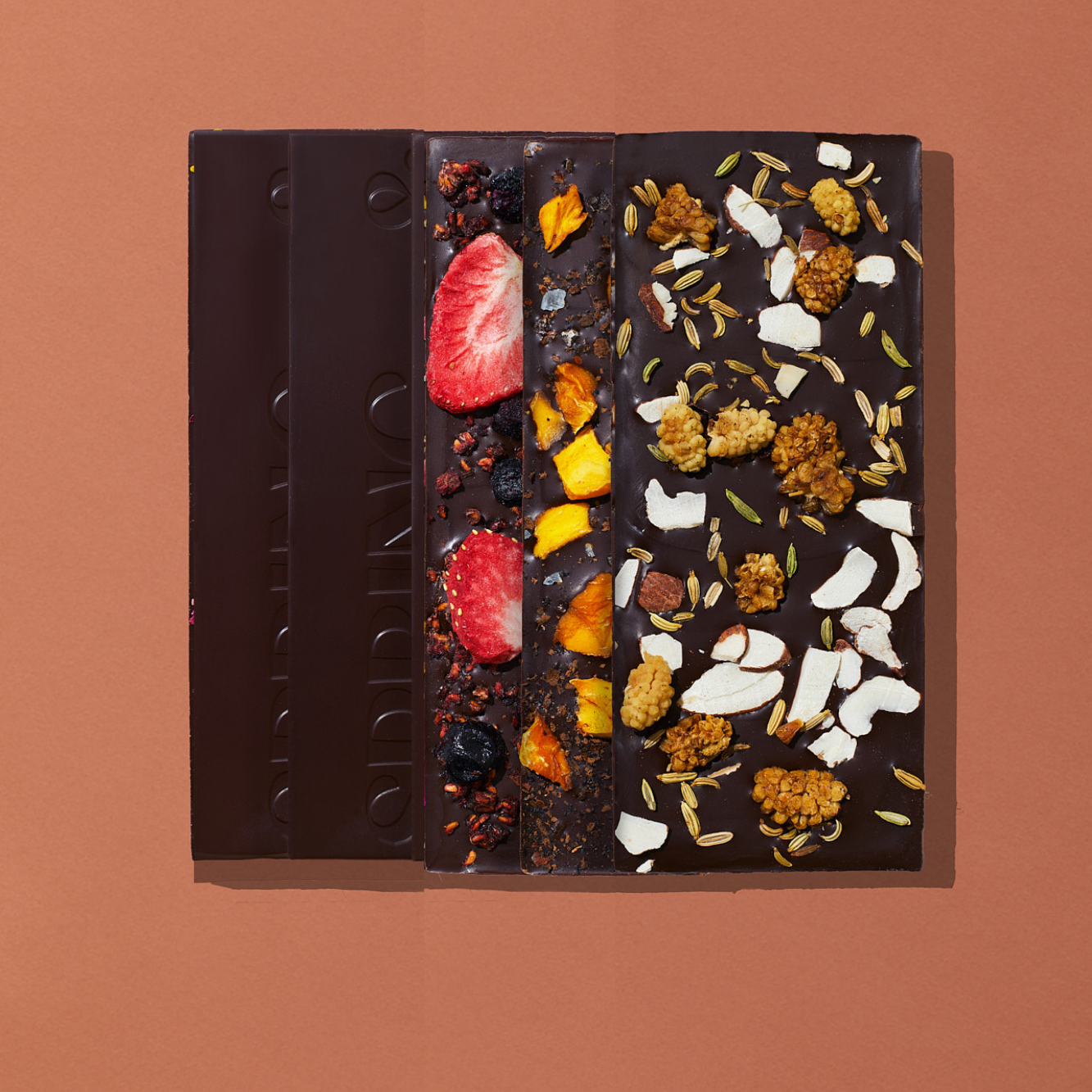 Five chocolate bars that are topped with fruits, florals, nuts, herbs and spices are laid out on a warm brown background.