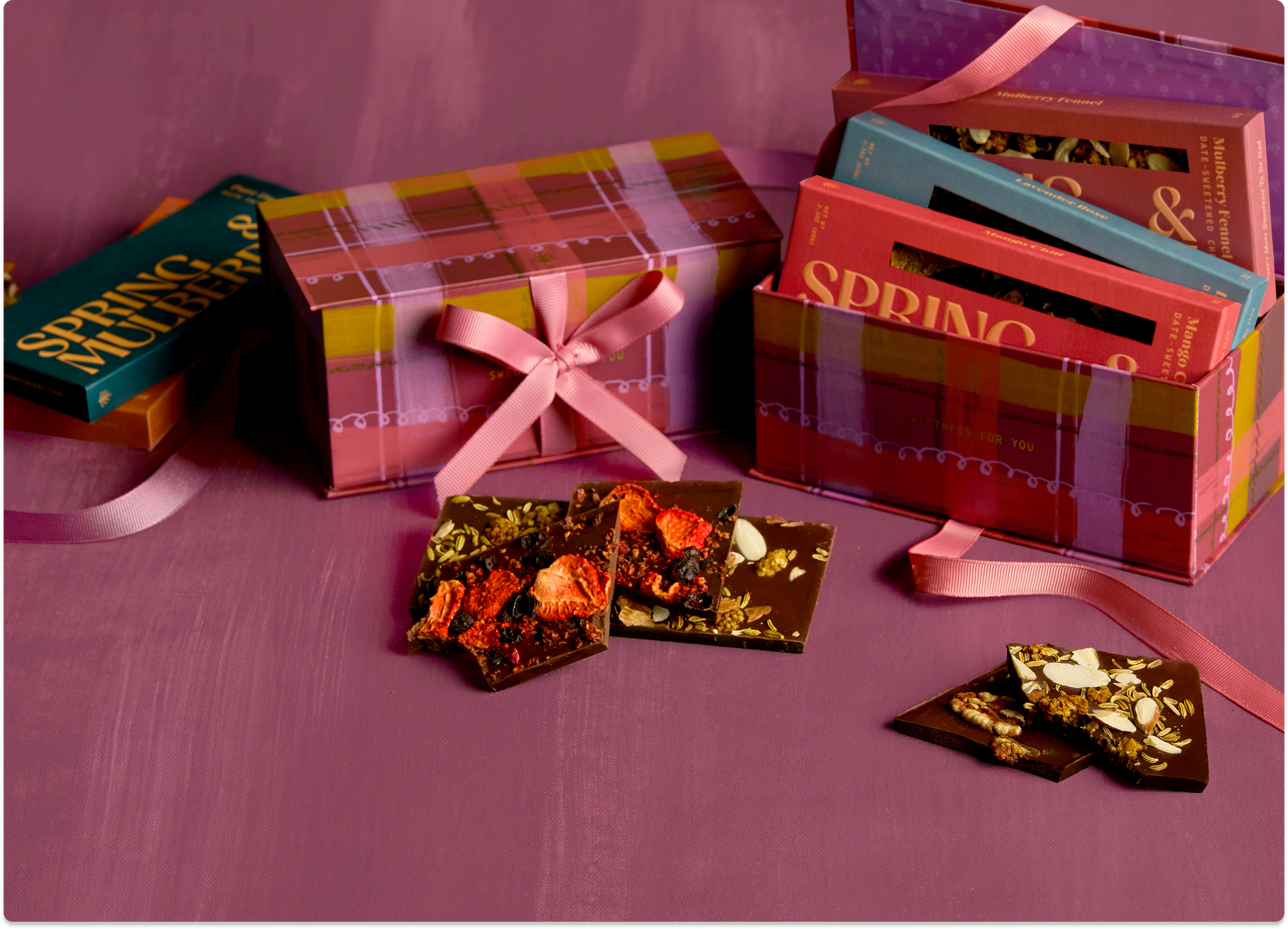 Two pink plaid gift boxes sit atop a painted pink background. One box is closed and tied with a ribbon. The other is open, showcasing Spring & Mulberry chocolate bars that sit inside.